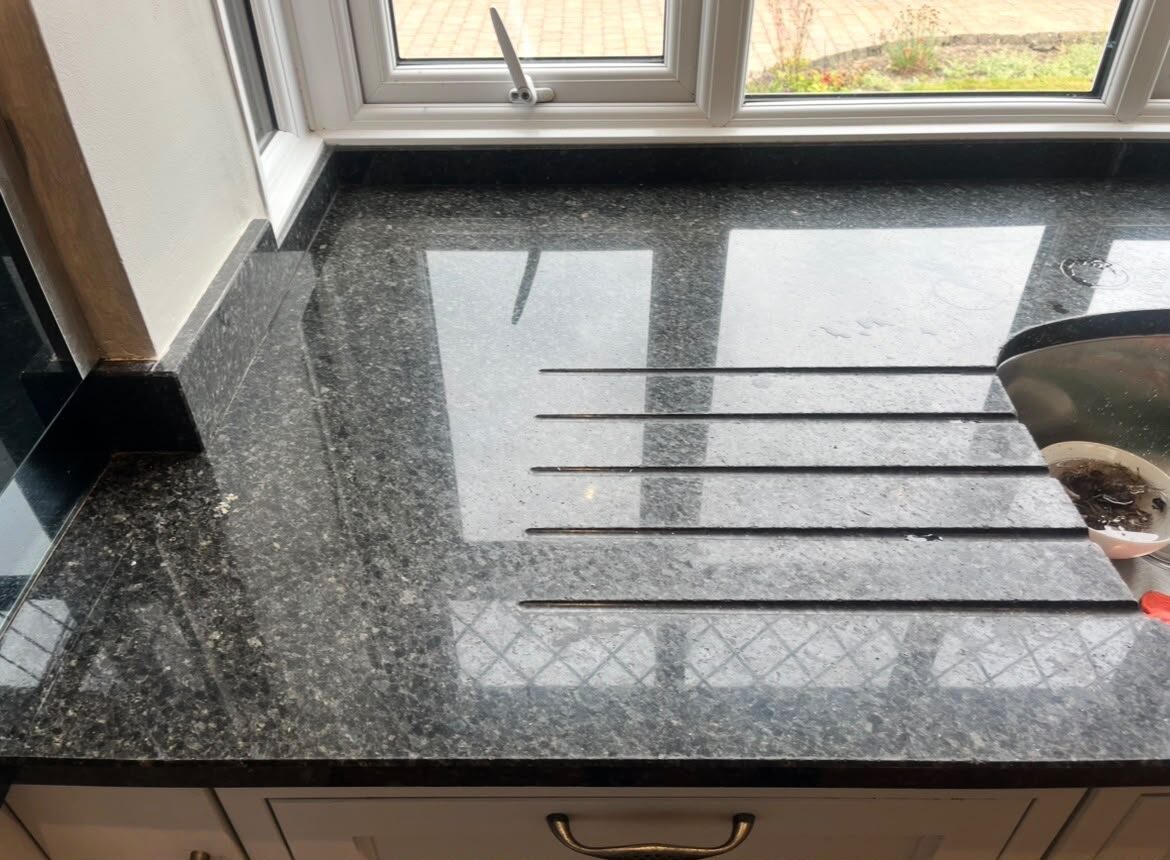 Grey granite worktop beside a sink with a dull, etched patch and limescale marks before restoration
